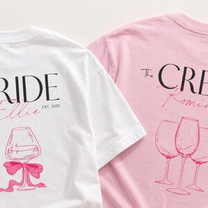 May include: A white t-shirt with black text reading The BRIDE, pink script reading Ellie, and a pink wine glass graphic with a bow, alongside a pink t-shirt with black text reading The CREW, pink script reading Romina, and three wine glass graphics.