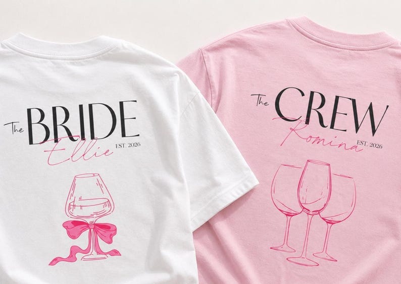 May include: A white t-shirt with black text reading The BRIDE, pink script reading Ellie, and a pink wine glass graphic with a bow, alongside a pink t-shirt with black text reading The CREW, pink script reading Romina, and three wine glass graphics.