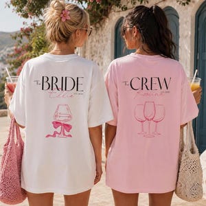 May include: A white t-shirt with the text The BRIDE, Ellie, and EST. 2026 above a wine glass graphic with a pink bow, and a light pink t-shirt with the text The CREW, Romina, and EST. 2026 above a graphic of three wine glasses in black and pink ink.