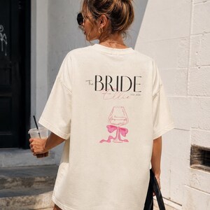 May include: An oversized cream-colored t-shirt featuring black text that reads The BRIDE, a pink script name Ellie, and the text EST. 2026. Below the text is a line-art illustration of a wine glass tied with a pink ribbon bow at the base.
