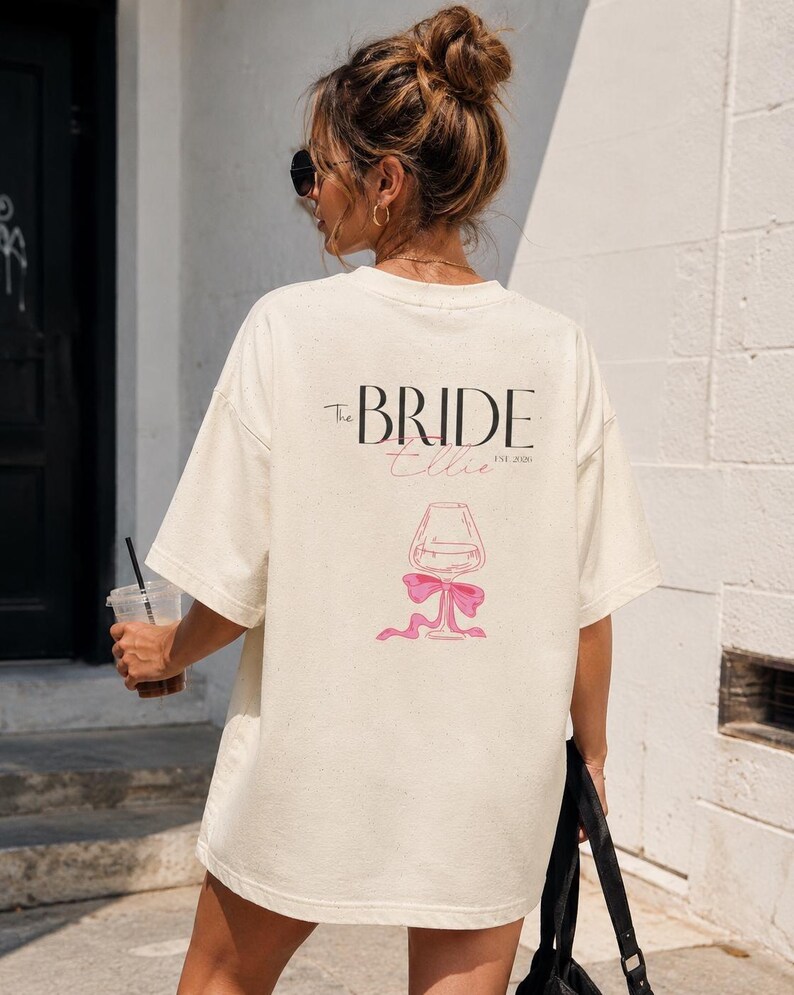 May include: An oversized cream-colored t-shirt featuring black text that reads The BRIDE, a pink script name Ellie, and the text EST. 2026. Below the text is a line-art illustration of a wine glass tied with a pink ribbon bow at the base.