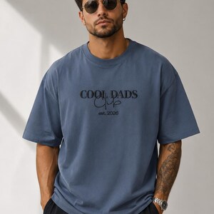 May include: A slate blue short-sleeved t-shirt featuring black embroidered text that reads COOL DADS Club and est. 2026. The shirt has a crew neckline and a relaxed fit, constructed from a solid-colored fabric with a smooth, matte finish.