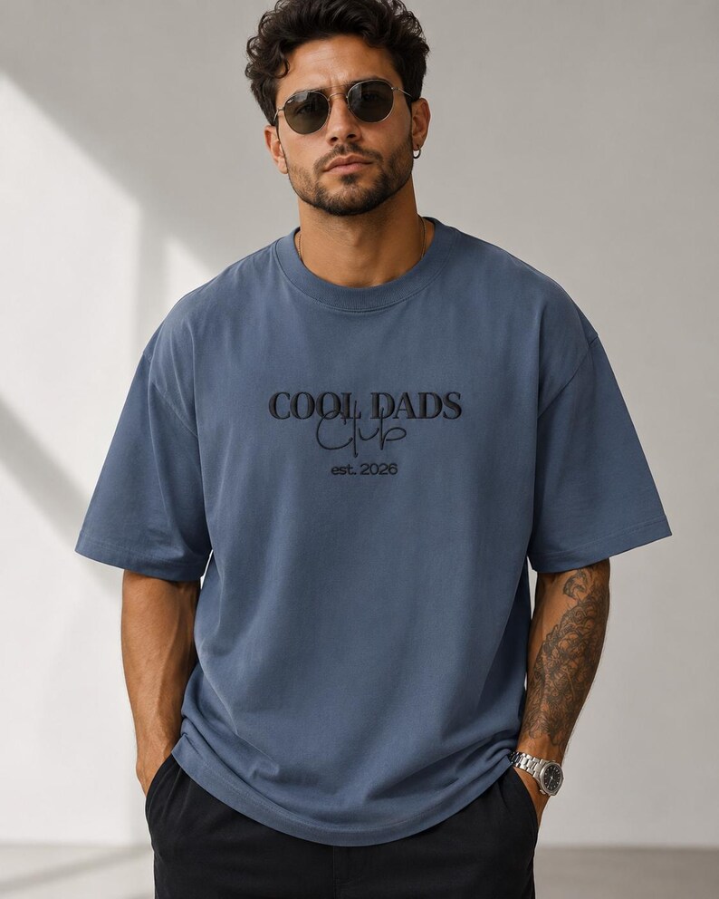 May include: A slate blue short-sleeved t-shirt featuring black embroidered text that reads COOL DADS Club and est. 2026. The shirt has a crew neckline and a relaxed fit, constructed from a solid-colored fabric with a smooth, matte finish.