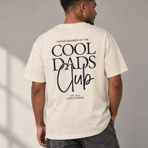 Könnte beinhalten: Ein übergroßes cremefarbenes T-Shirt mit schwarzem Text auf der Rückseite, der PROUD MEMBER OF THE COOL DADS Club EST. 2023 LIAM & EMMY lautet. Das T-Shirt hat kurze Ärmel und einen Rundhalsausschnitt und wird mit einer dunkelgrauen Jeans kombiniert.