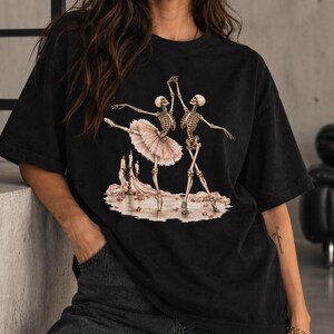 Pu&ograve; includere: Una t-shirt nera con una stampa grafica di due scheletri che ballano. Uno scheletro indossa un tut&ugrave; rosa pallido. La scena include un candelabro con candele accese e petali di rosa sparsi sul terreno, creando un'illustrazione molto dettagliata.