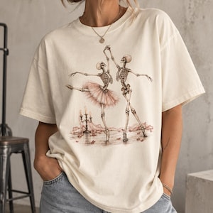 Pu&ograve; includere: Una t-shirt a maniche corte color bianco sporco con una stampa grafica di due scheletri che ballano. Uno scheletro indossa un tut&ugrave; rosa, mentre l'altro &egrave; nudo. La scena include un candelabro con candele accese e petali rosa sparsi.