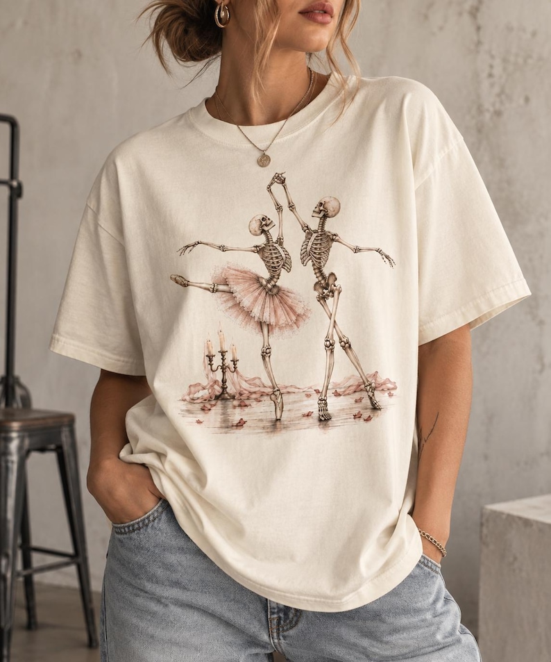 Pu&ograve; includere: Una t-shirt a maniche corte color bianco sporco con una stampa grafica di due scheletri che ballano. Uno scheletro indossa un tut&ugrave; rosa, mentre l'altro &egrave; nudo. La scena include un candelabro con candele accese e petali rosa sparsi.