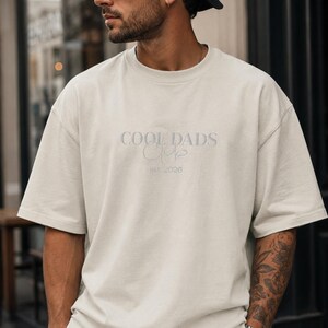 May include: An off-white, short-sleeved crew neck t-shirt featuring the embroidered text COOL DADS Club and est. 2026 in a matching light tone on the chest. The garment has a relaxed, loose fit with a standard hemline and a simple, clean design.