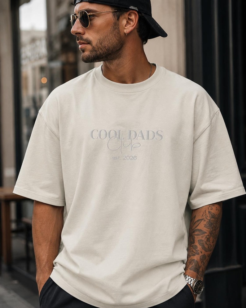 May include: An off-white, short-sleeved crew neck t-shirt featuring the embroidered text COOL DADS Club and est. 2026 in a matching light tone on the chest. The garment has a relaxed, loose fit with a standard hemline and a simple, clean design.