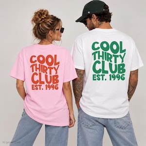 Può includere: Una t-shirt rosa chiaro e una t-shirt bianca, entrambe con la scritta COOL THIRTY CLUB EST. 1996 stampata sul retro. La maglietta rosa presenta lettere rosse, mentre quella bianca ha lettere verdi. Entrambe sono abbinate a jeans in denim blu.
