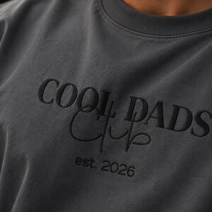 May include: A charcoal gray crewneck sweatshirt featuring black embroidery on the chest. The text reads COOL DADS in a bold serif font, with the word Club written in a cursive script underneath, followed by the text est. 2026 in a smaller font.