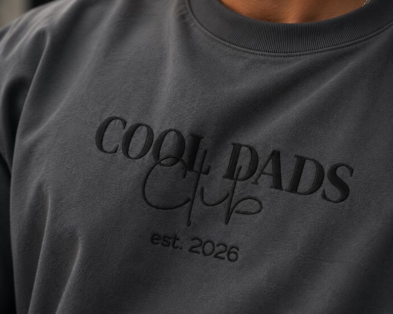 May include: A charcoal gray crewneck sweatshirt featuring black embroidery on the chest. The text reads COOL DADS in a bold serif font, with the word Club written in a cursive script underneath, followed by the text est. 2026 in a smaller font.