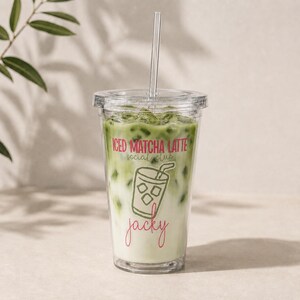 Op de afbeelding: Een doorzichtige plastic beker gevuld met een ijs-matcha latte, voorzien van een deksel en een recht rietje. De beker toont de tekst ICED MATCHA LATTE social club in roze en groen, een lijntekening van een drankje en de naam Jacky in roze.