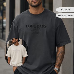 May include: A charcoal gray short-sleeved t-shirt featuring the embroidered text COOL DADS CLUB and est. 2026 in black thread. A circular inset shows the same t-shirt design in a light beige color. The shirt has a crew neckline and loose fit.