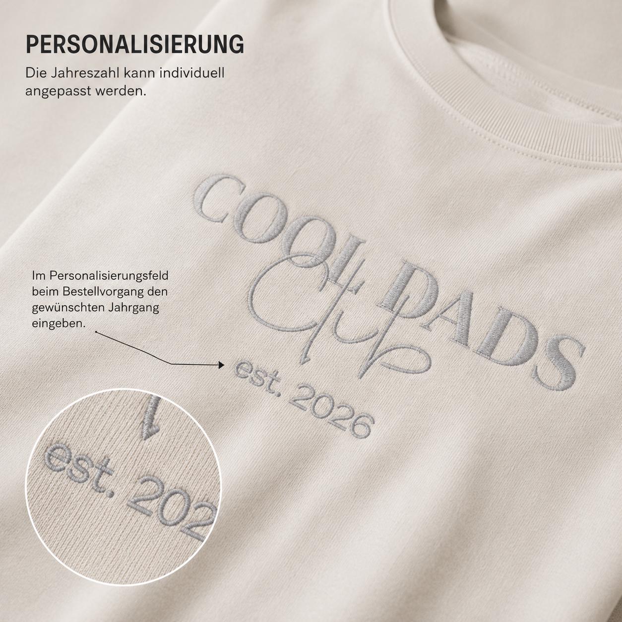 May include: A beige sweatshirt featuring the embroidered text COOL DADS CLUB and est. 2026 in a grey thread. The image includes German text regarding personalization options, with a circular inset showing a close-up of the embroidered year detail.