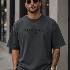 May include: A charcoal gray short-sleeve t-shirt featuring the text COOL DADS CLUB in a bold serif font with the word Club in a cursive script underneath and the text est. 2026 printed below in a smaller, simple sans-serif font.