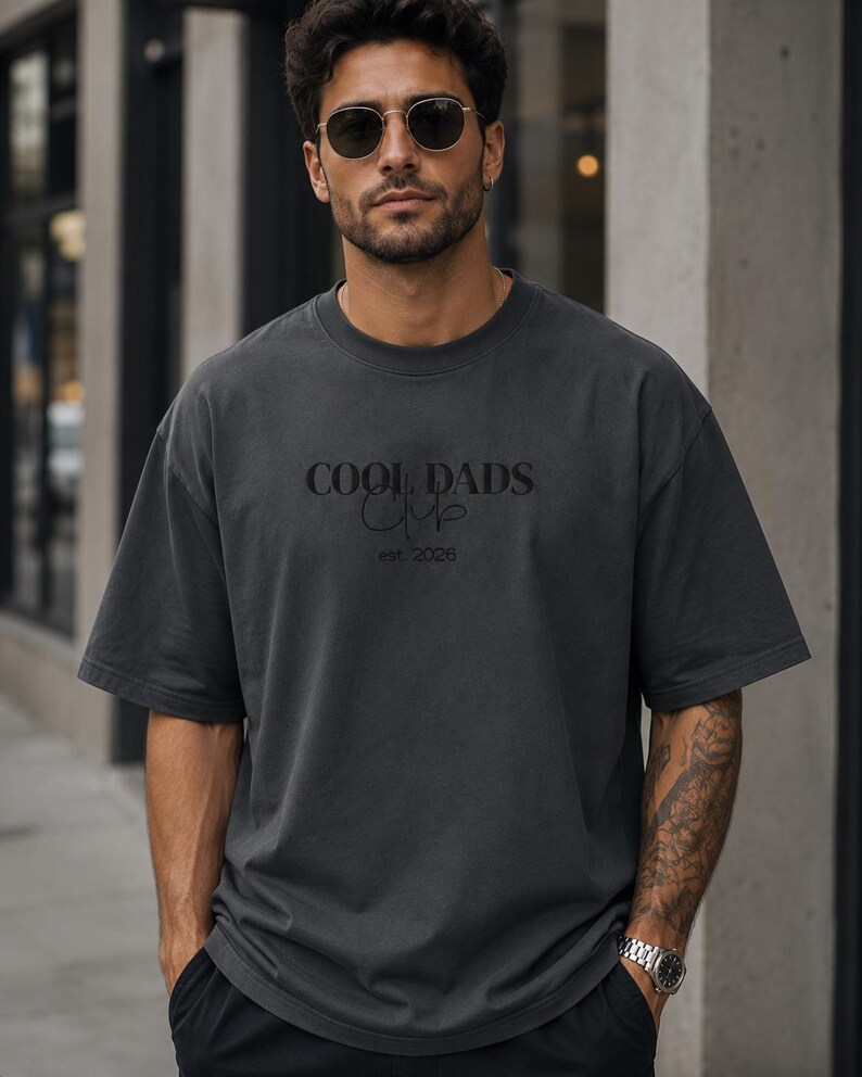 May include: A charcoal gray short-sleeve t-shirt featuring the text COOL DADS CLUB in a bold serif font with the word Club in a cursive script underneath and the text est. 2026 printed below in a smaller, simple sans-serif font.