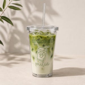 Könnte beinhalten: Ein durchsichtiger Kunststoffbecher gefüllt mit einem Eismatcha-Latte, inklusive Deckel und transparentem Strohhalm. Der Becher zeigt den grünen Schriftzug ICED MATCHA LATTE, eine Strichzeichnung eines Getränks und den Namen emilia in Schreibschrift.