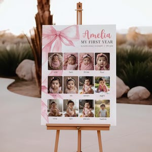 May include: A wooden easel displays a pink and white sign with the name "Amelia" and photos of a baby. The sign has a pink bow graphic and photos of the baby from one to twelve months old. The easel is made of light brown wood.
