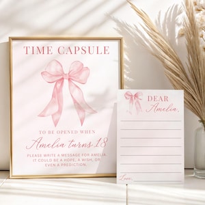 May include: A framed pink time capsule sign and matching note card. The sign features a watercolor bow and text that reads "Time Capsule" and "To be opened when Amelia turns 18." The note card says "Dear Amelia" and has lines for writing.