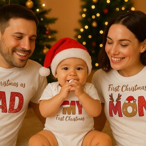 Könnte beinhalten: Eine dreiköpfige Familie trägt passende weiße T-Shirts und einen Baby-Body für ihr erstes Weihnachten. Auf den Shirts steht „First Christmas as Dad“, „First Christmas as Mom“ und „First Christmas as My“. Das Baby trägt einen Weihnachtsmannhut.