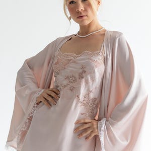May include: A pale pink satin lingerie set, including a slip dress with lace detailing and a matching robe. The robe has wide sleeves with white lace trim. The slip dress features a square neckline and lace accents. A pearl necklace completes the ensemble.