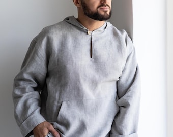May include: A light gray long-sleeved hooded shirt featuring a single button closure at the neckline, a front kangaroo pocket, and cuffed sleeves. The garment is constructed from a textured fabric and includes matching gray trousers.