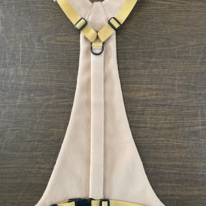 Op de afbeelding: Een beige fleece hondenharnas met gele banden en zwarte gespen. Het harnas heeft een D-ring aan de achterkant om een ​​riem aan te bevestigen.