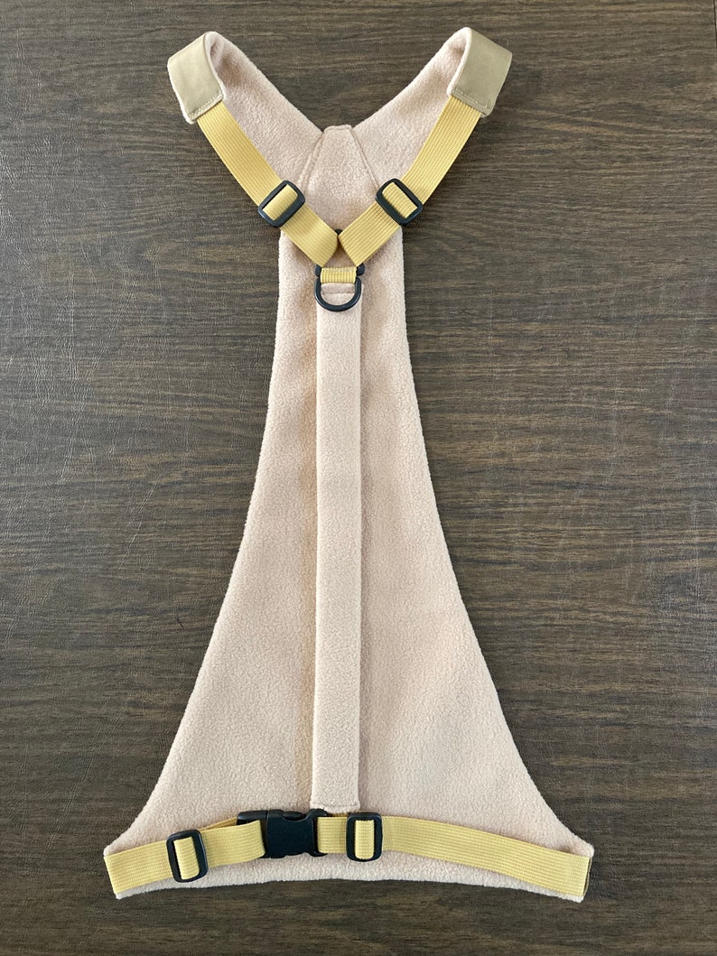 Op de afbeelding: Een beige fleece hondenharnas met gele banden en zwarte gespen. Het harnas heeft een D-ring aan de achterkant om een ​​riem aan te bevestigen.