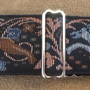 Puede incluir: Un collar negro para perros con un dise&ntilde;o tejido que presenta perros corriendo en marr&oacute;n y azul, junto con patrones florales. El collar tiene herrajes met&aacute;licos plateados.