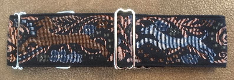 Puede incluir: Un collar negro para perros con un dise&ntilde;o tejido que presenta perros corriendo en marr&oacute;n y azul, junto con patrones florales. El collar tiene herrajes met&aacute;licos plateados.