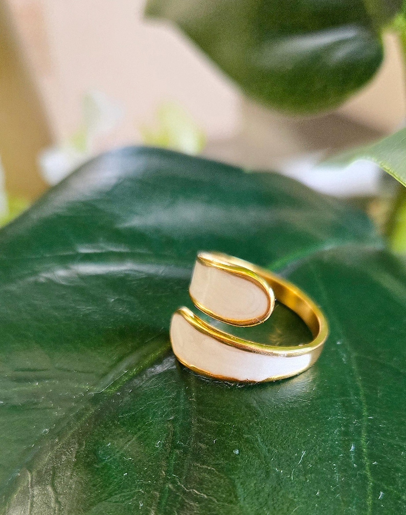 Puede incluir: Un anillo dorado con incrustaciones de esmalte blanco. El anillo tiene un dise&ntilde;o abierto y envolvente, con las secciones de esmalte blanco curv&aacute;ndose alrededor del dedo. El anillo est&aacute; sobre una hoja verde.