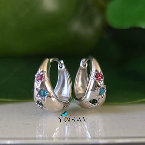 May include: A pair of silver hoop earrings with star-shaped accents. Each earring features a red, blue, and green gemstone. The earrings are displayed on a reflective surface with a green background. The brand name "YOSAV Jewels" is visible.