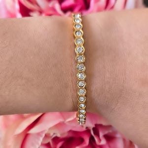 May include: A gold bracelet with clear, round gemstones set in a bezel design. The bracelet is worn on a wrist, with pink flowers in the background. The bracelet is a delicate piece of jewelry.