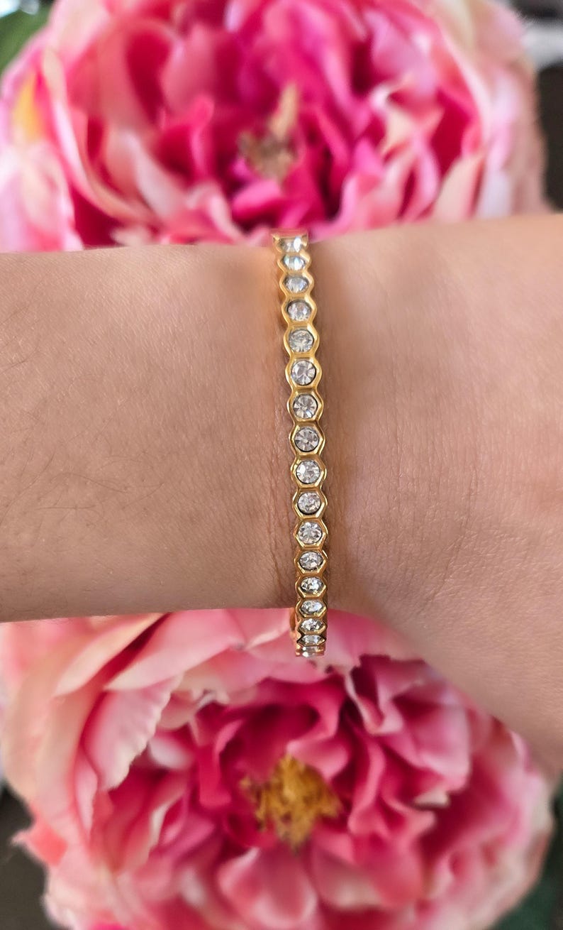 May include: A gold bracelet with clear, round gemstones set in a bezel design. The bracelet is worn on a wrist, with pink flowers in the background. The bracelet is a delicate piece of jewelry.