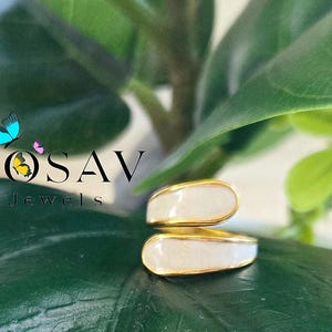 Puede incluir: Un anillo dorado con una incrustaci&oacute;n de esmalte blanco. El anillo tiene un dise&ntilde;o abierto y superpuesto. La imagen muestra el anillo sobre un fondo de hojas verdes. El texto "YOSAV Jewels" es visible.