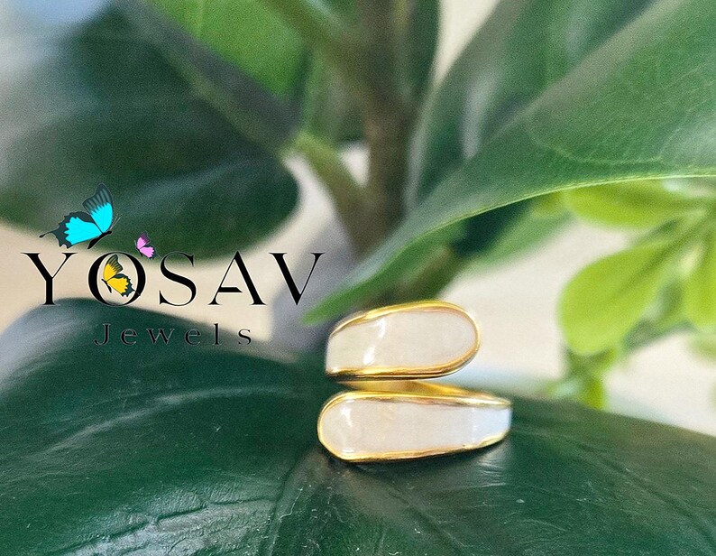 Puede incluir: Un anillo dorado con una incrustaci&oacute;n de esmalte blanco. El anillo tiene un dise&ntilde;o abierto y superpuesto. La imagen muestra el anillo sobre un fondo de hojas verdes. El texto "YOSAV Jewels" es visible.