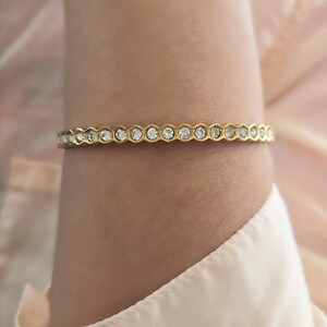May include: A gold-toned bracelet with a repeating pattern of clear, round gemstones. The bracelet is worn on a wrist, and the background is a soft, neutral color. The bracelet's design is elegant and suitable for various occasions.
