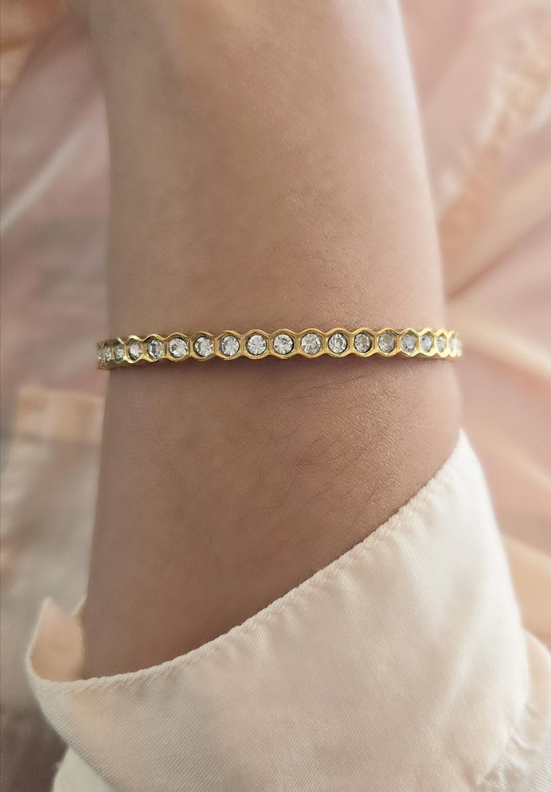 May include: A gold-toned bracelet with a repeating pattern of clear, round gemstones. The bracelet is worn on a wrist, and the background is a soft, neutral color. The bracelet's design is elegant and suitable for various occasions.