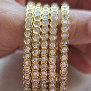 May include: Six gold-colored rings with a honeycomb design, each cell containing a clear, round gemstone. The rings are stacked on a finger, showcasing the intricate detail and sparkle of the jewelry. The rings are a warm gold color.