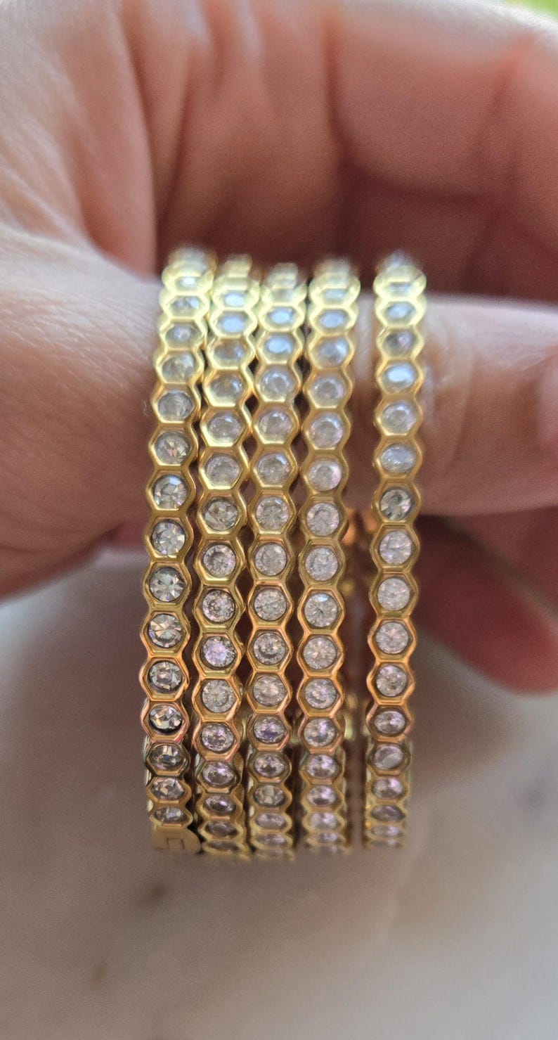 May include: Six gold-colored rings with a honeycomb design, each cell containing a clear, round gemstone. The rings are stacked on a finger, showcasing the intricate detail and sparkle of the jewelry. The rings are a warm gold color.