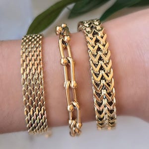 May include: Three gold-colored bracelets are displayed on a wrist. The bracelets feature different designs: a woven chain, a chain of connected links with spheres, and a chevron pattern. The bracelets are made of a shiny, metallic material.