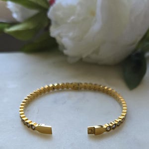 May include: Gold-toned bracelet with a row of small, clear gemstones. The open cuff design features a rectangular clasp. The bracelet is displayed on a white surface with a blurred background of white flowers and green leaves.