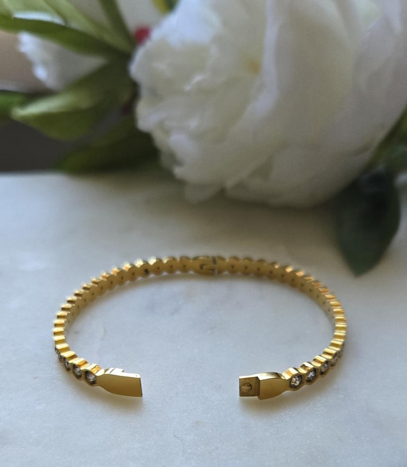 May include: Gold-toned bracelet with a row of small, clear gemstones. The open cuff design features a rectangular clasp. The bracelet is displayed on a white surface with a blurred background of white flowers and green leaves.