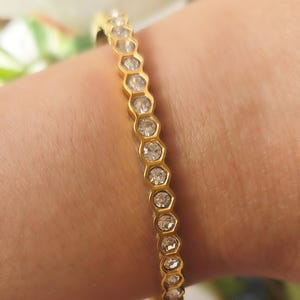 May include: A gold-colored bracelet with a honeycomb design, featuring clear, round gemstones set in each cell. The bracelet is worn on a wrist, showcasing its delicate and elegant appearance. The gemstones sparkle, reflecting light.