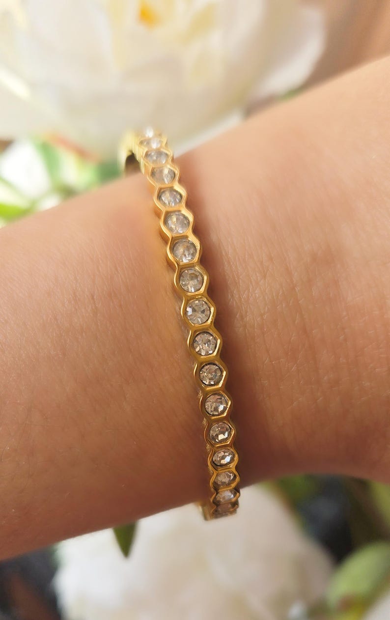 May include: A gold-colored bracelet with a honeycomb design, featuring clear, round gemstones set in each cell. The bracelet is worn on a wrist, showcasing its delicate and elegant appearance. The gemstones sparkle, reflecting light.