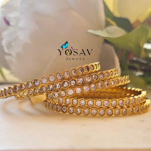 May include: Four gold bangles with a honeycomb design, each cell set with a clear gemstone. The bangles are stacked, with the logo "YOSAV Jewels" visible in the background. The jewelry is displayed on a white surface.