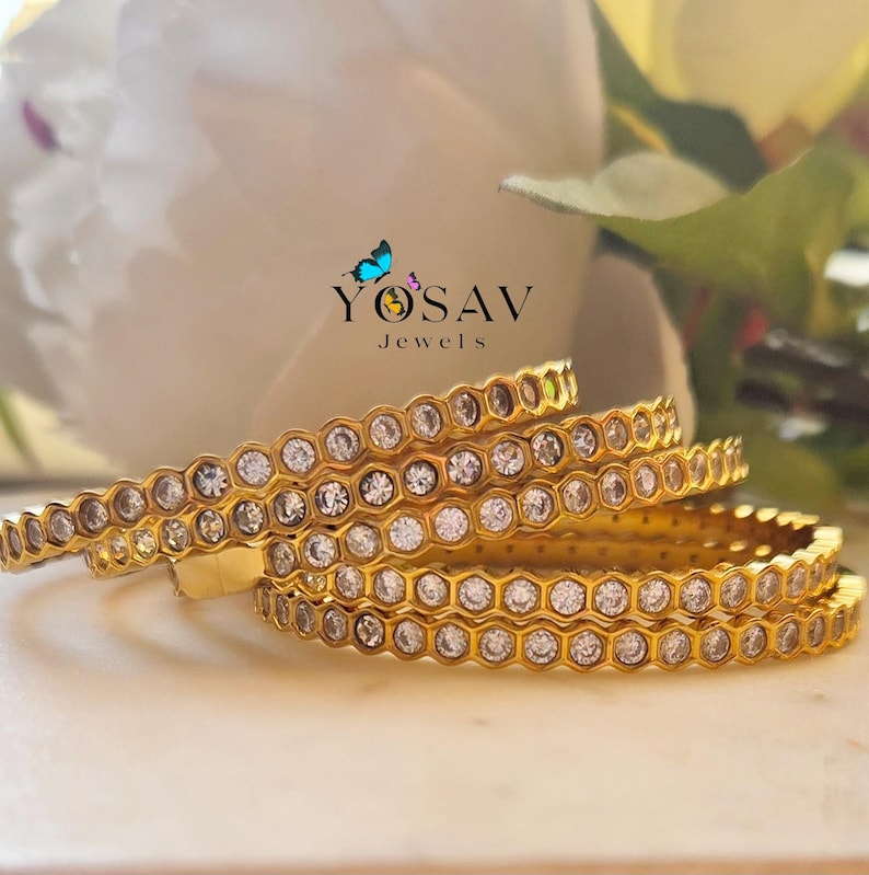 May include: Four gold bangles with a honeycomb design, each cell set with a clear gemstone. The bangles are stacked, with the logo "YOSAV Jewels" visible in the background. The jewelry is displayed on a white surface.