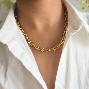 May include: A gold-toned chain necklace featuring a series of elongated, U-shaped links connected by small, spherical beads. The necklace rests against the collar of a white button-down shirt, showcasing a polished, metallic finish throughout the design.
