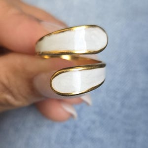 Puede incluir: Un anillo dorado con un dise&ntilde;o de esmalte blanco. El anillo se envuelve alrededor del dedo con dos secciones ovaladas de esmalte blanco, cada una delineada con oro. La pieza de joyer&iacute;a es un dise&ntilde;o moderno y minimalista.